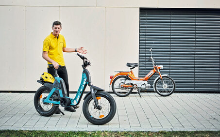 auto touring-Redakteur Alex Fischer steht zwischen zwei Zweirädern. Links befindet sich das moderne, blaue Elektrofahrrad, die Yamaha Booser easy, mit einem gelben Helm auf dem Gepäckträger. Rechts steht das klassische, orangefarbene Moped – die Puch Maxi.