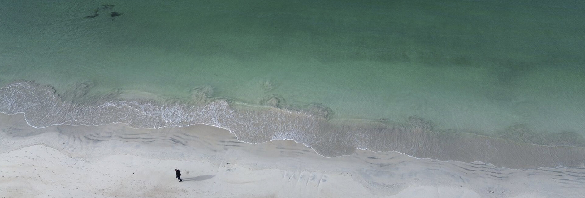 Drohnenfoto: Aufnahme vom Strand und dem türkisgrünem Meer, eine Person spaziert im Sand.