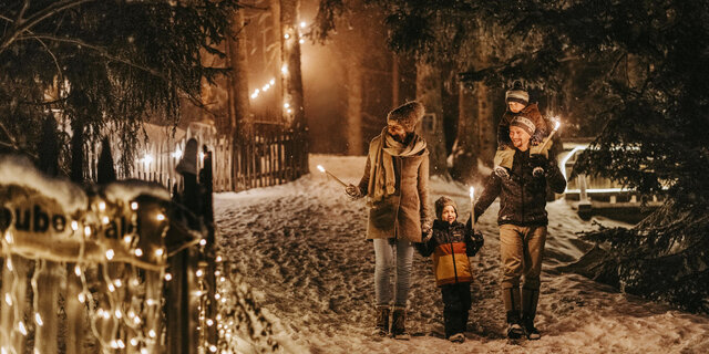 Eltern gehen mit zwei Kindern im Schnee im Wald. Die Werg ist mit beleuchtet.