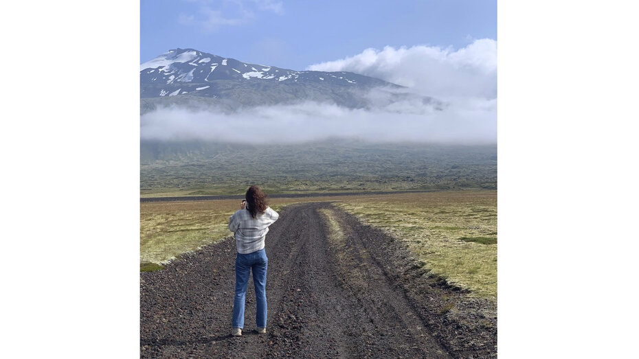 Eine Frau steht auf einer Schotterstraße in Island, man sieht sie von hinten und sie fotografiert die Landschaft.