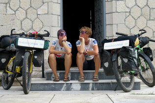 Zwei Frauen sitzen auf den Stufen vor einer offenen Tür, jeweils neben ihnen ihre Fahrräder mit Gepäck. 