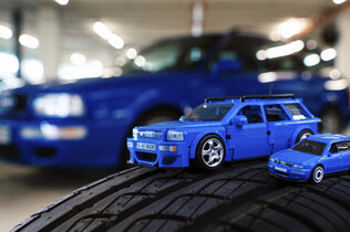 Audi-Modell aus dem Mattel Brick Shop von oben. Daneben steht das gleiche Modell von Hot Wheels. Beide stehen auf einem echten Reifen, im Hintergrund der original Audi..