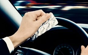 Das Bild zeigt die Hände von einem Autofahrer im Fahrzeug. Er sitzt am Steuer und hält mit einer Hand das Lenkrad, während er in der linken Hand ein Tablettenblister hält. 