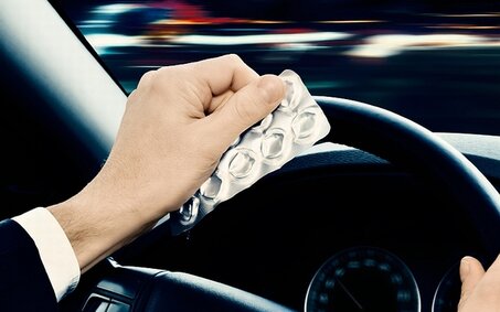 Das Bild zeigt die Hände von einem Autofahrer im Fahrzeug. Er sitzt am Steuer und hält mit einer Hand das Lenkrad, während er in der linken Hand ein Tablettenblister hält. 