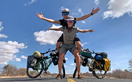 Die beiden Radabenteurerinnen Tanja Willers und Johanna Hochedlinger auf einer Straße in Sambia. Tanja sitzt auf dem Rücken von Johanna, beide stehen vor ihren Rädern.
