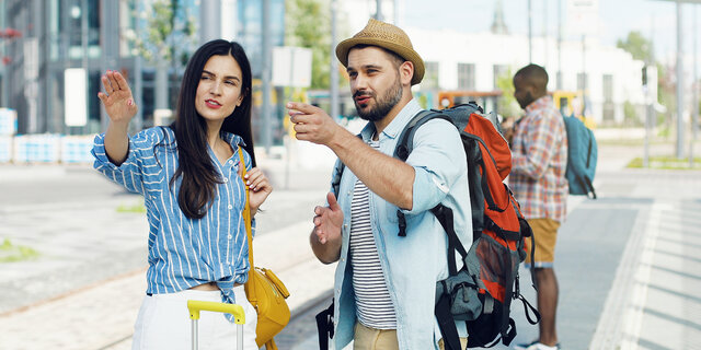 Caucasian young beautiful woman tourist standing at bus stop with suitcase. Handsome male traveller with backpack coming to ask the route. Man asking for right way Girl explaining how to get somewhere