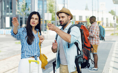 Caucasian young beautiful woman tourist standing at bus stop with suitcase. Handsome male traveller with backpack coming to ask the route. Man asking for right way Girl explaining how to get somewhere