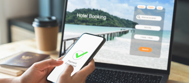 Hand vor einem Laptop mit offener Website zur Hotelbuchung für den Urlaub. Handy zeigt als Nachricht "abgeschlossene Zahlung" an.