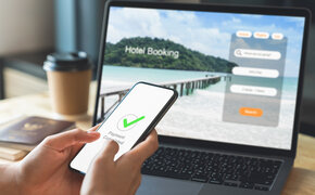 Hand vor einem Laptop mit offener Website zur Hotelbuchung für den Urlaub. Handy zeigt als Nachricht "abgeschlossene Zahlung" an.