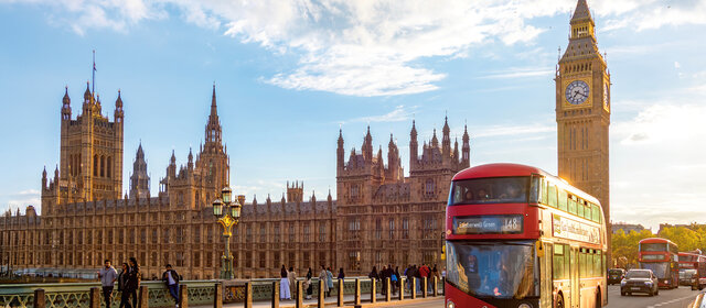 Blick auf Big Ben von einer Brücke aus, es ist sonnig. Es fährt ein roter Bus vorbei.