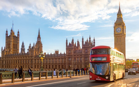 Blick auf Big Ben von einer Brücke aus, es ist sonnig. Es fährt ein roter Bus vorbei.