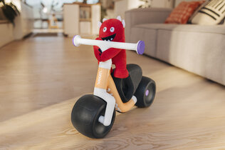 Imagebild des neuen Woom Wow Laufrads mit kleinem roten Stoffmonster am Lenker, im Hintergrund ein Wohnzimmer.