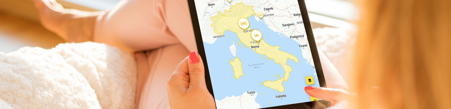 Eine Person sitzt entspannt und benutzt ein Tablet mit der ÖAMTC-App, um eine Karte von Italien zu betrachten.