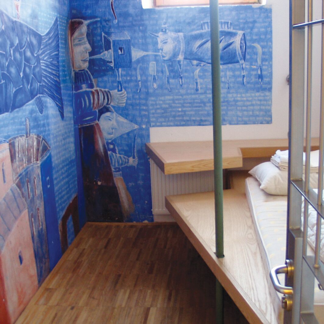 Ein Zimmer mit Wandgemälde im blauen Farbton, einem schmalen Bett und Holzmöbeln.