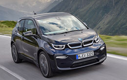BMW i3 E-Auto in dunkelblau fährt auf einer Straße, im Hintergrund Wiese und Berge.