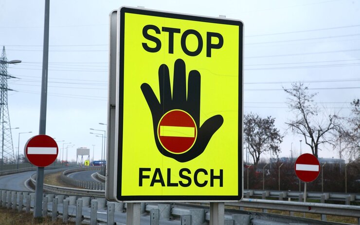 An einer Autobahnauffahrt steht ein großes neongelbes STOP FALSCH Schild. Eine schwarze Hand hat an der Handfläche ein Einfahrt verboten Schild. Im Hintergrund sind zwei weitere Schilder mit der Bedeutung Einfahrt verboten sichtbar.