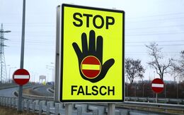 An einer Autobahnauffahrt steht ein großes neongelbes STOP FALSCH Schild. Eine schwarze Hand hat an der Handfläche ein Einfahrt verboten Schild. Im Hintergrund sind zwei weitere Schilder mit der Bedeutung Einfahrt verboten sichtbar.