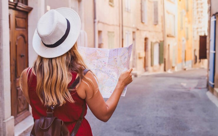 Eine Frau in einem roten Kleid und Hut betrachtet eine Landkarte in einer engen, sonnigen Gasse.