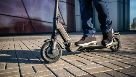 Nahaufnahme von Füßen, die auf einem Scooter stehen. Die Schuhe sind sportlich und der Scooter hat ein modernes Design.