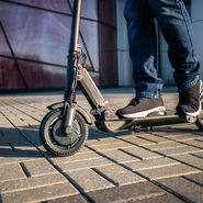 Nahaufnahme von Füßen, die auf einem Scooter stehen. Die Schuhe sind sportlich und der Scooter hat ein modernes Design.