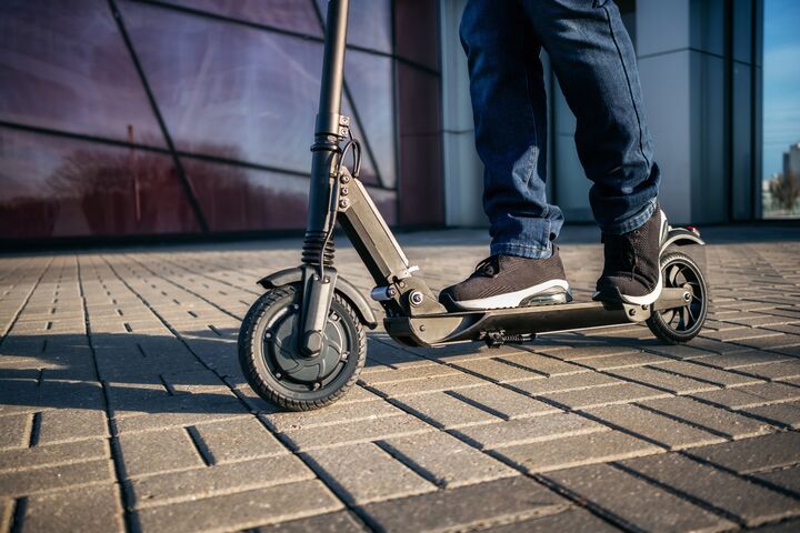 Nahaufnahme von Füßen, die auf einem Scooter stehen. Die Schuhe sind sportlich und der Scooter hat ein modernes Design.