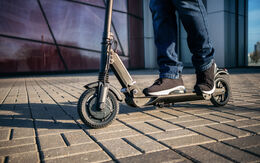Nahaufnahme von Füßen, die auf einem Scooter stehen. Die Schuhe sind sportlich und der Scooter hat ein modernes Design.