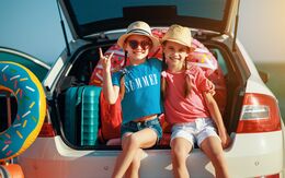 Zwei Mädchen sitzen lächelnd im Kofferraum eines vollgepackten Autos, bereit für den Urlaub. Neben und hinter ihnen befindet sich noch ein aufblasbarer Schwimmreifen.