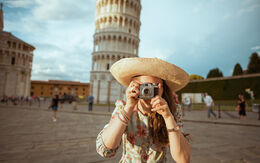 Eine Frau mit Strohhut macht ein Foto mit einer Kamera vor dem schiefen Turm von Pisa.