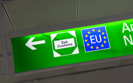 Ein grünes Schild mit einem weißen Pfeil nach links. Auf dem Schild sind zwei Symbole abgebildet: links ein weißes Sechseck mit der Aufschrift "Zoll Customs"  und rechts ein blaues Quadrat mit gelben Sternen, dass die Flagge der EU darstellt.