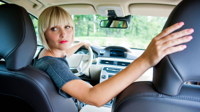 Eine Frau mit blonden Haaren sitzt im Auto am Steuer und schaut über die rechte Schulter nach hinten. Sie trägt ein graues T-Shirt. Der rechte Arm liegt auf der Beifahrersitzlehne.