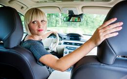 Eine Frau mit blonden Haaren sitzt im Auto am Steuer und schaut über die rechte Schulter nach hinten. Sie trägt ein graues T-Shirt. Der rechte Arm liegt auf der Beifahrersitzlehne.