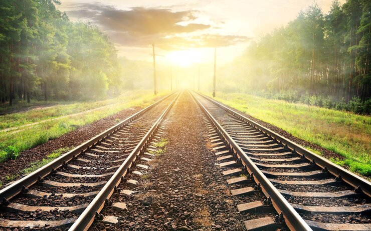 Zwei Eisenbahnschienen verlaufen parallel durch eine grüne Landschaft in Richtung eines Sonnenaufgangs oder Sonnenuntergangs am Horizont.