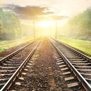 Zwei Eisenbahnschienen verlaufen parallel durch eine grüne Landschaft in Richtung eines Sonnenaufgangs oder Sonnenuntergangs am Horizont.