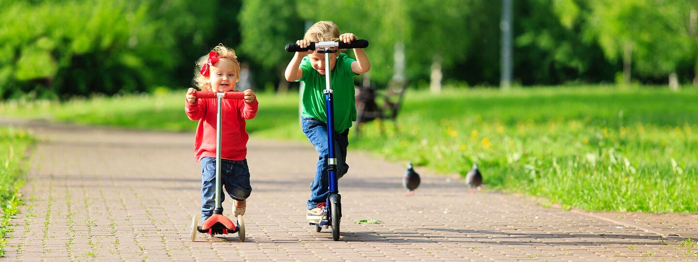 Ein kleines Mädchen und ein kleiner Junge fahren mit dem Tretroller bzw. Micro-Scooter auf einem Gehsteig im Park.
