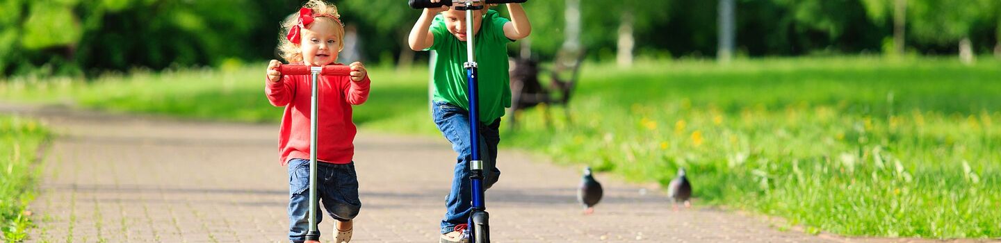 Ein kleines Mädchen und ein kleiner Junge fahren mit dem Tretroller bzw. Micro-Scooter auf einem Gehsteig im Park.
