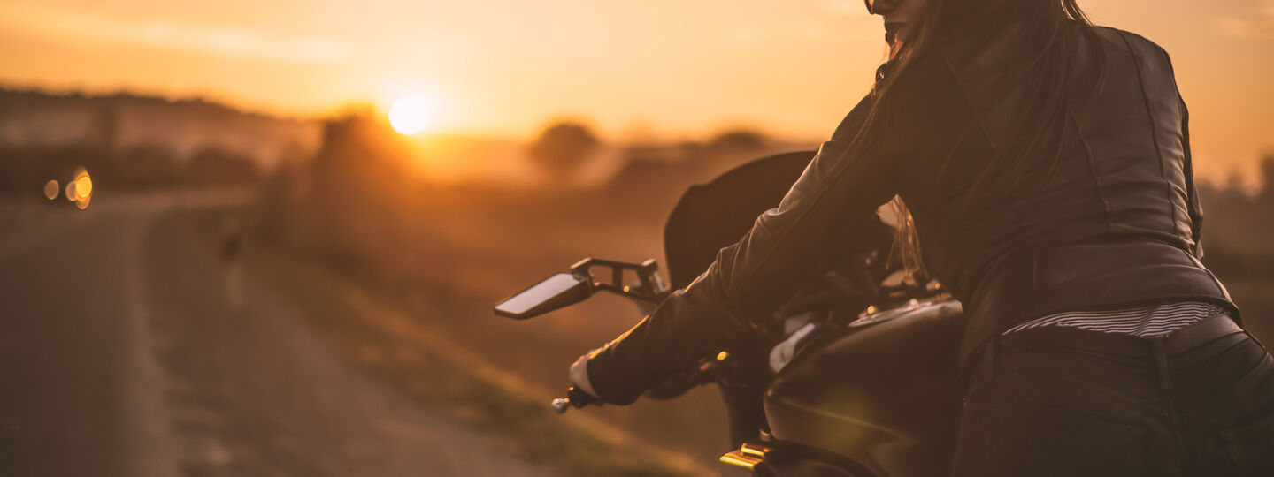 Eine Frau mit Sonnenbrille und langen, braunen Haaren, trägt eine schwarze Lederjacke und schwarze Hose. Sie sitzt auf einem Motorrad und blickt zur Seite. Im Vordergrund befindet sich eine Landstraße, die von einer ländlichen Umgebung und einem Sonnenuntergang umrahmt wird.