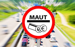 Ein Verkehrszeichen zeigt ein Symbol, das für die Maut steht. Im Hintergrund sieht man eine mehrspurige Autobahn mit Fahrzeugen.