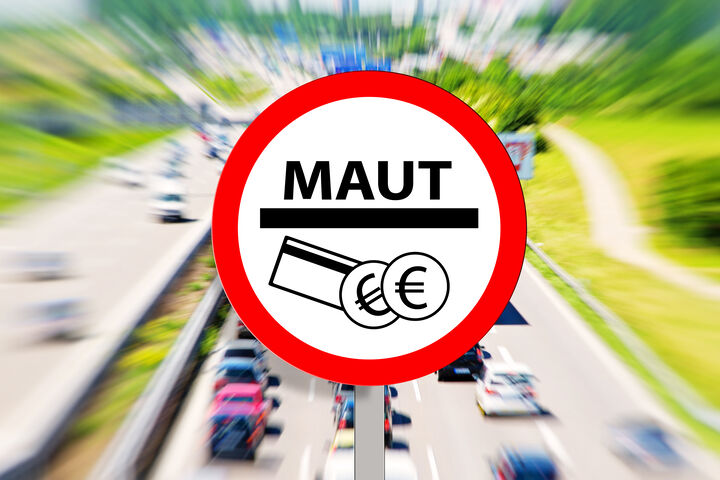 Ein Verkehrszeichen zeigt ein Symbol, das für die Maut steht. Im Hintergrund sieht man eine mehrspurige Autobahn mit Fahrzeugen.