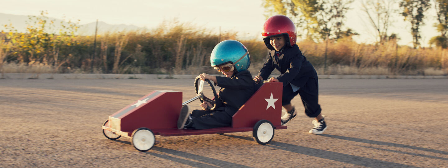 Zwei Kinder mit großen Helmen haben Spaß mit einem lustigen Raketenfahrzeug. Ein Kind mit blauem Helm sitzt im Fahrzeug, das zweite Kind mit rotem Helm schiebt hinten das Gefährt an.