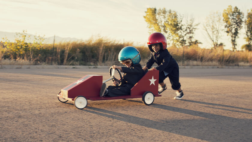 Zwei Kinder mit großen Helmen haben Spaß mit einem lustigen Raketenfahrzeug. Ein Kind mit blauem Helm sitzt im Fahrzeug, das zweite Kind mit rotem Helm schiebt hinten das Gefährt an.