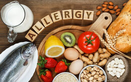 Auf einem Holztisch befinden sich ein Fisch, eine Kanne Milch, Erdbeeren, Nüsse, ein Brot, Eier, eine Kiwi, eine Tomate, Bohnen, eine Zitrone und Sesam. Über dem Brett bilden Holzbuchstaben das Wort "ALLERGY".