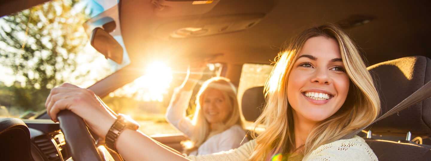 Junge Frau am Steuer im Auto bei sonnigem Wetter unterwegs. Sie schaut nach links und lacht. Am Beifahrersitz befindet sich eine weitere Frau.