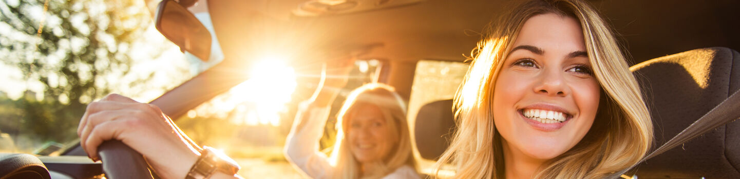 Junge Frau am Steuer im Auto bei sonnigem Wetter unterwegs. Sie schaut nach links und lacht. Am Beifahrersitz befindet sich eine weitere Frau.