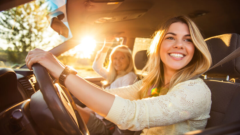 Junge Frau am Steuer im Auto bei sonnigem Wetter unterwegs. Sie schaut nach links und lacht. Am Beifahrersitz befindet sich eine weitere Frau.