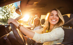 Junge Frau am Steuer im Auto bei sonnigem Wetter unterwegs. Sie schaut nach links und lacht. Am Beifahrersitz befindet sich eine weitere Frau.