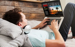 Ein junger Mann liegt im Bett, trägt Kopfhörer um den Hals und schaut auf einem Laptop eine Streaming-Serie.