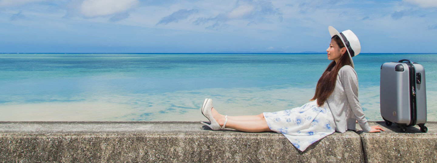 Eine junge Frau in Sommerkleidung und mit Sonnenhut sitzt auf einer kleinen Mauer neben dem Meer, neben ihr steht ein Koffer.