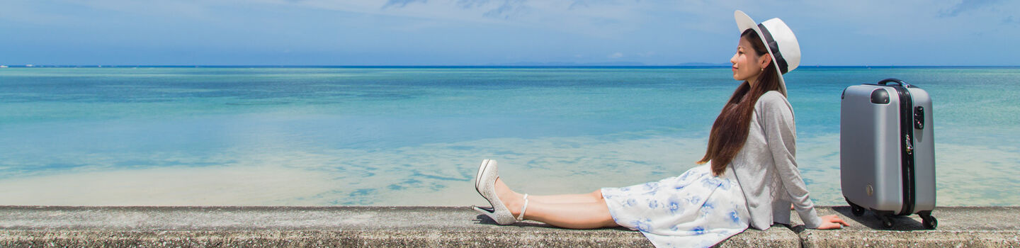 Eine junge Frau in Sommerkleidung und mit Sonnenhut sitzt auf einer kleinen Mauer neben dem Meer, neben ihr steht ein Koffer.