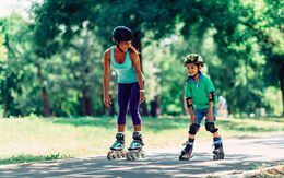 Mutter und Sohn beim Rollschuhlaufen bzw. Inlineskaten im Park. Beide tragen Helm, der Junge auch Knie- und Ellbogenschützer.