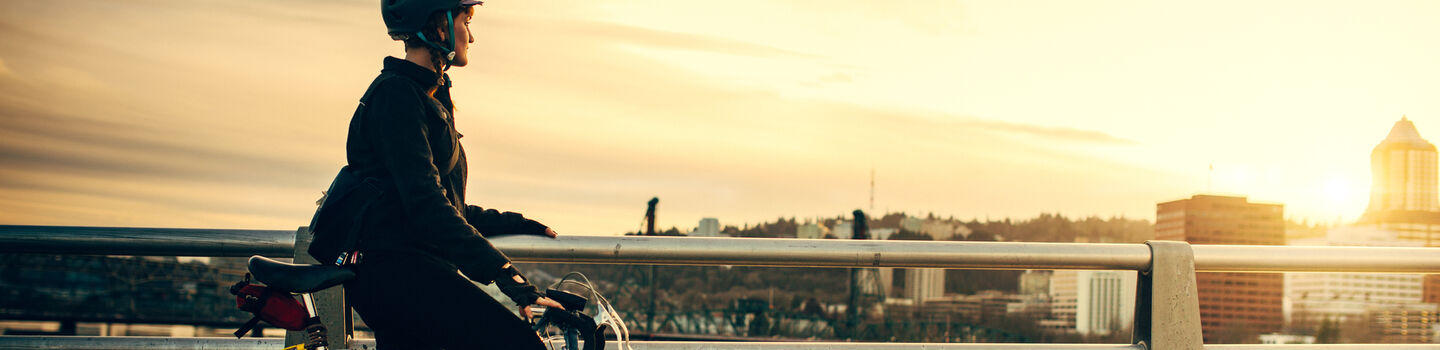 Radfahrerin mit Helm steht auf Weg mit Fahrrad und blickt auf einen Fluss bei Sonnenuntergang.